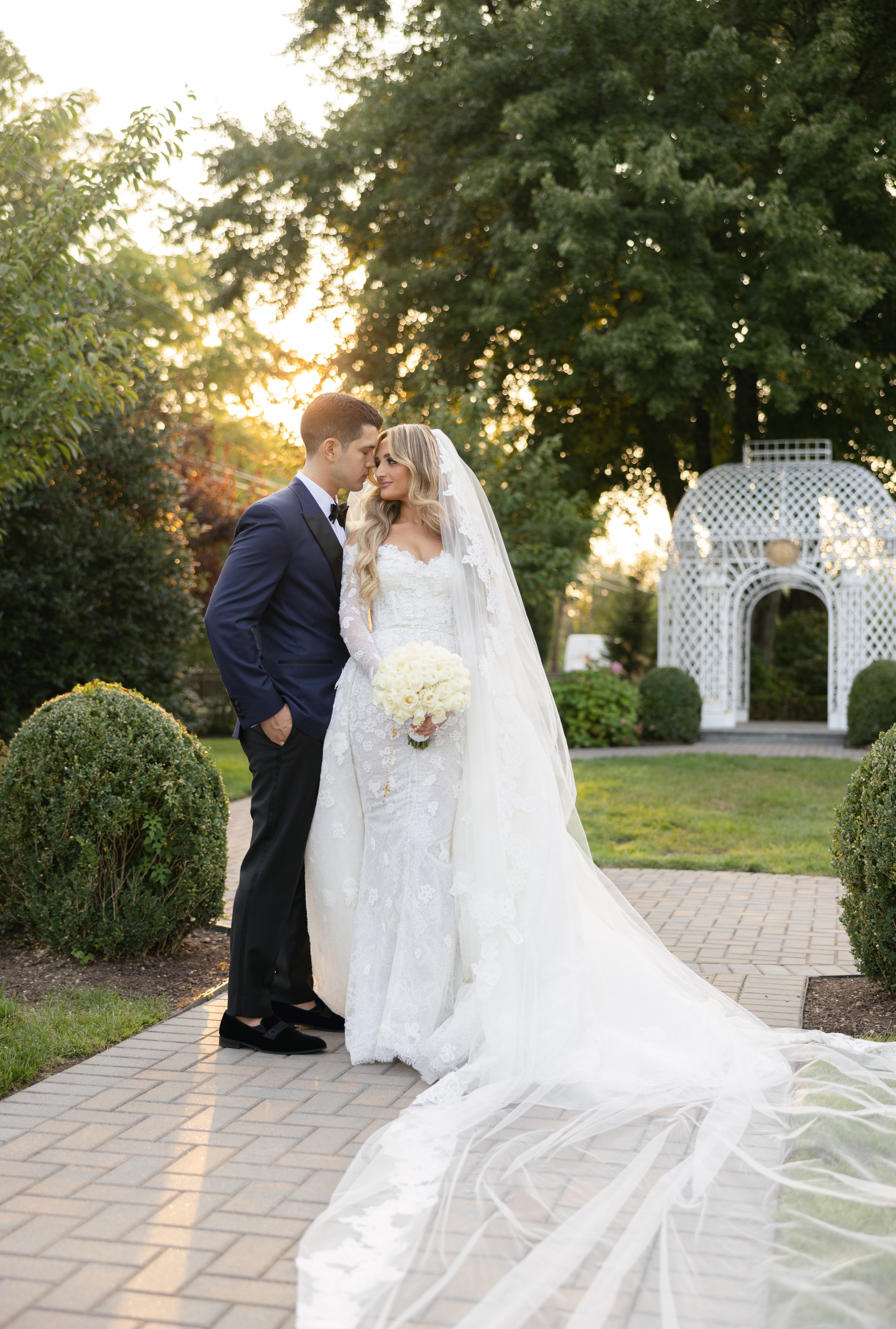 Bride with long veil and bouquet beside groom on a garden walkway
